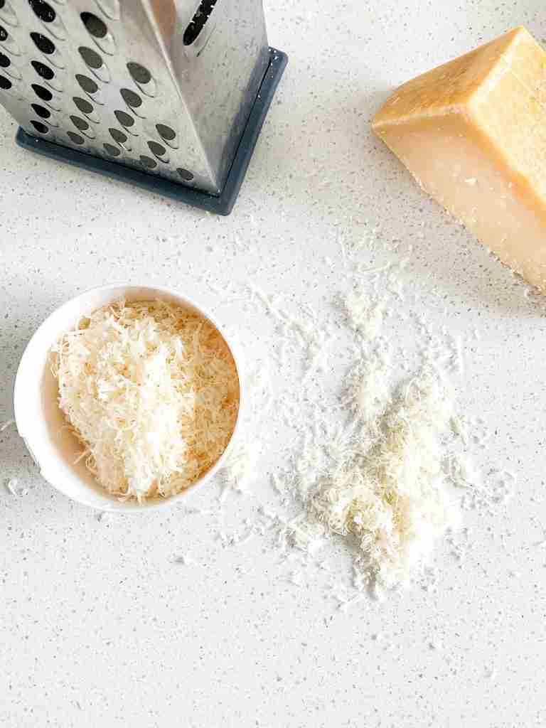 shredded cheese in a cup with a cheese grater beside it and a hunk of cheese