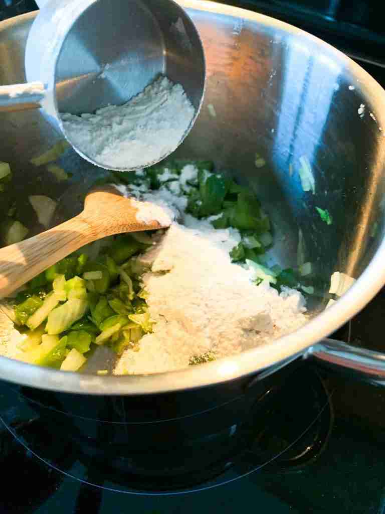 flour being added into a pan full of veggies for a roux