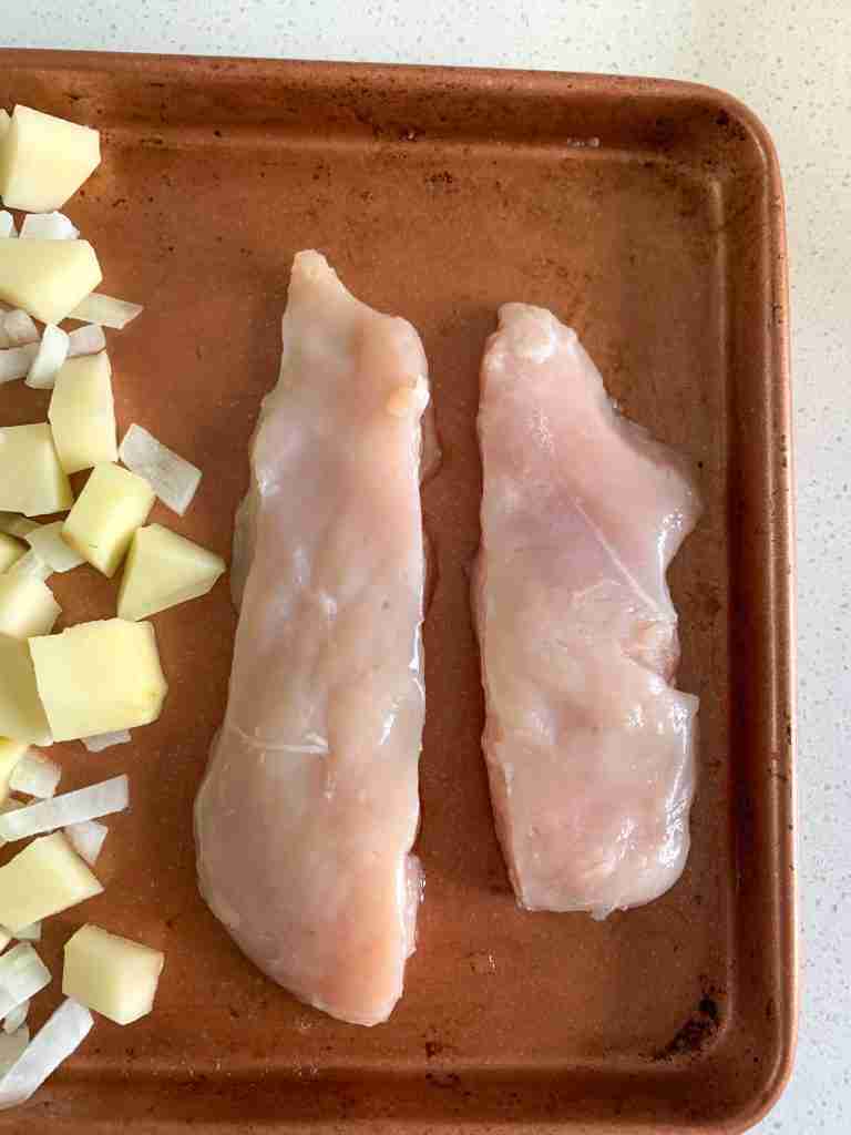 raw chicken placed on a sheet pan next to raw potatoes