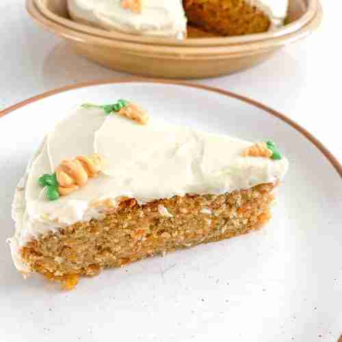 slice of cake on a plate in front of the full cake with little carrot decorations on top