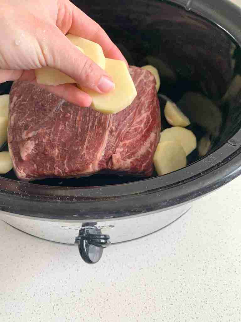 hand placing potatoes around roast in a crockpot