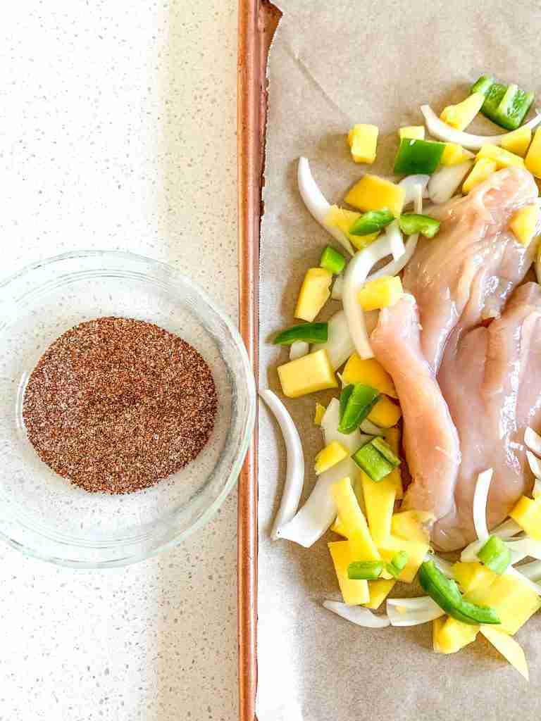 raw chicken and veggies on a pan with a bowl of seasoning beside it