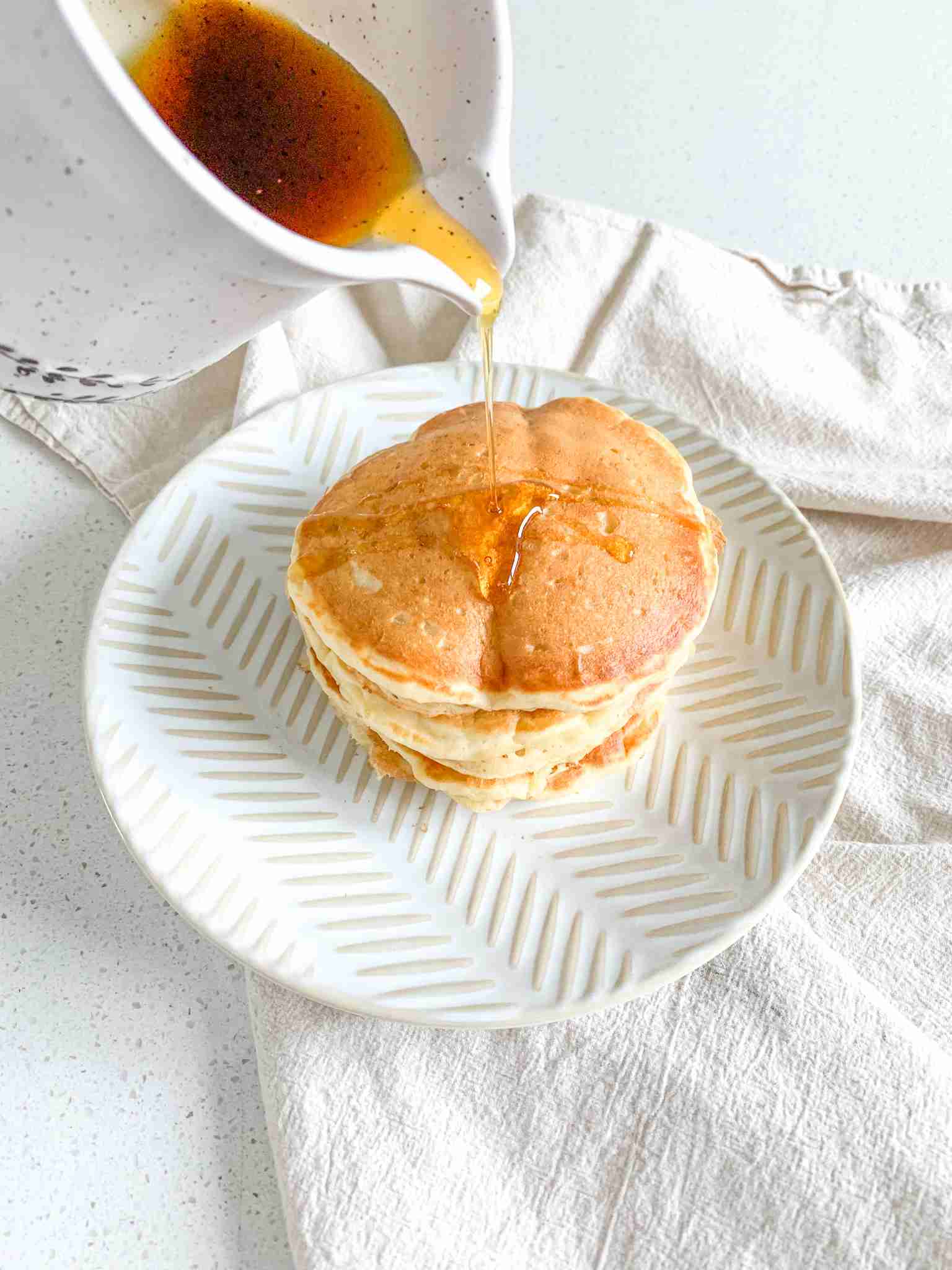 maple syrup getting poured over a freshly made stack of sourdough buttermilk pancakes