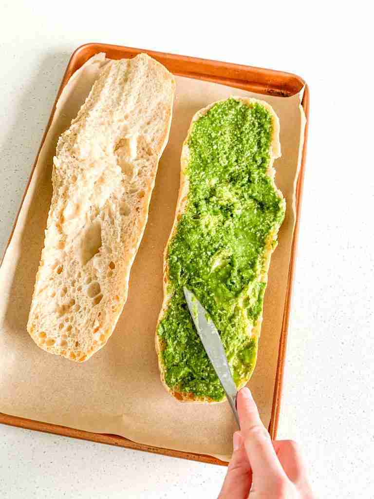 pesto being spread on French bread for sourdough pesto garlic bread