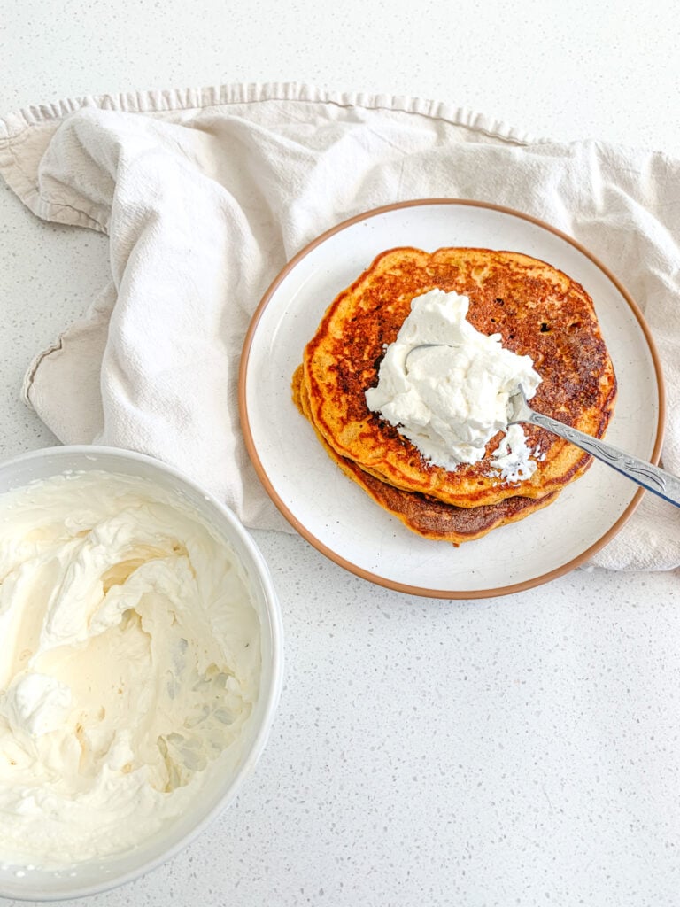 topping stack of pumpkin pancakes with whipped cream