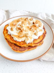 a stack of sourdough pumpkin pancakes freshly made and topped with whipped cream, nuts, and syrup
